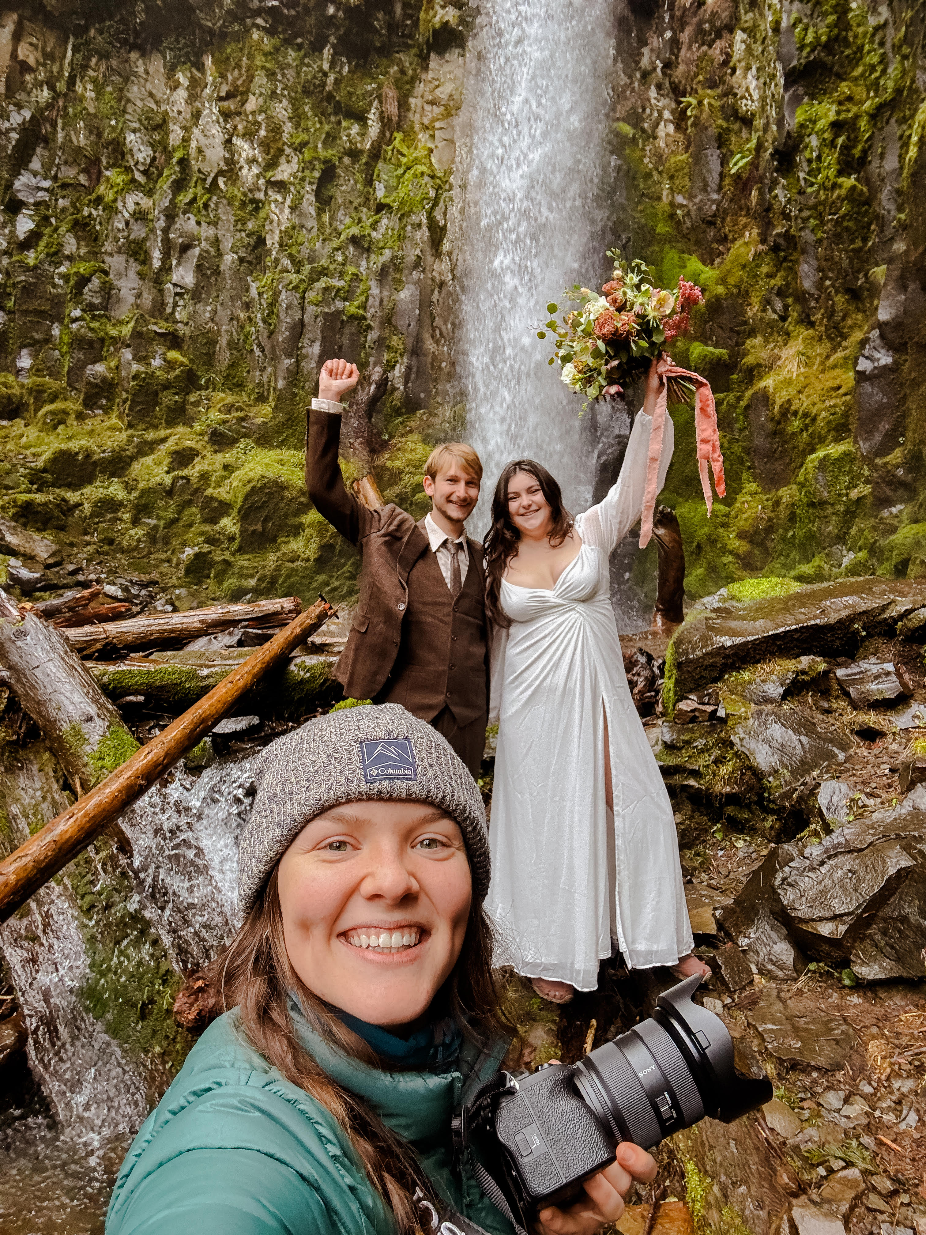 A photographer takes a selfie with a smiling couple in wedding attire standing in front of a waterfall, surrounded by lush greenery.